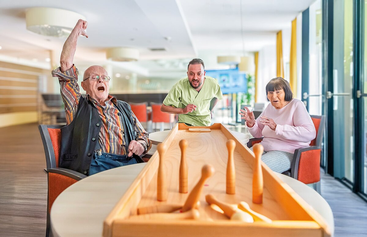Ein älterer Mann jubelt mit erhobenem Arm, während er mit einer älteren Frau und einem jüngeren Mann Tischkegeln spielt.