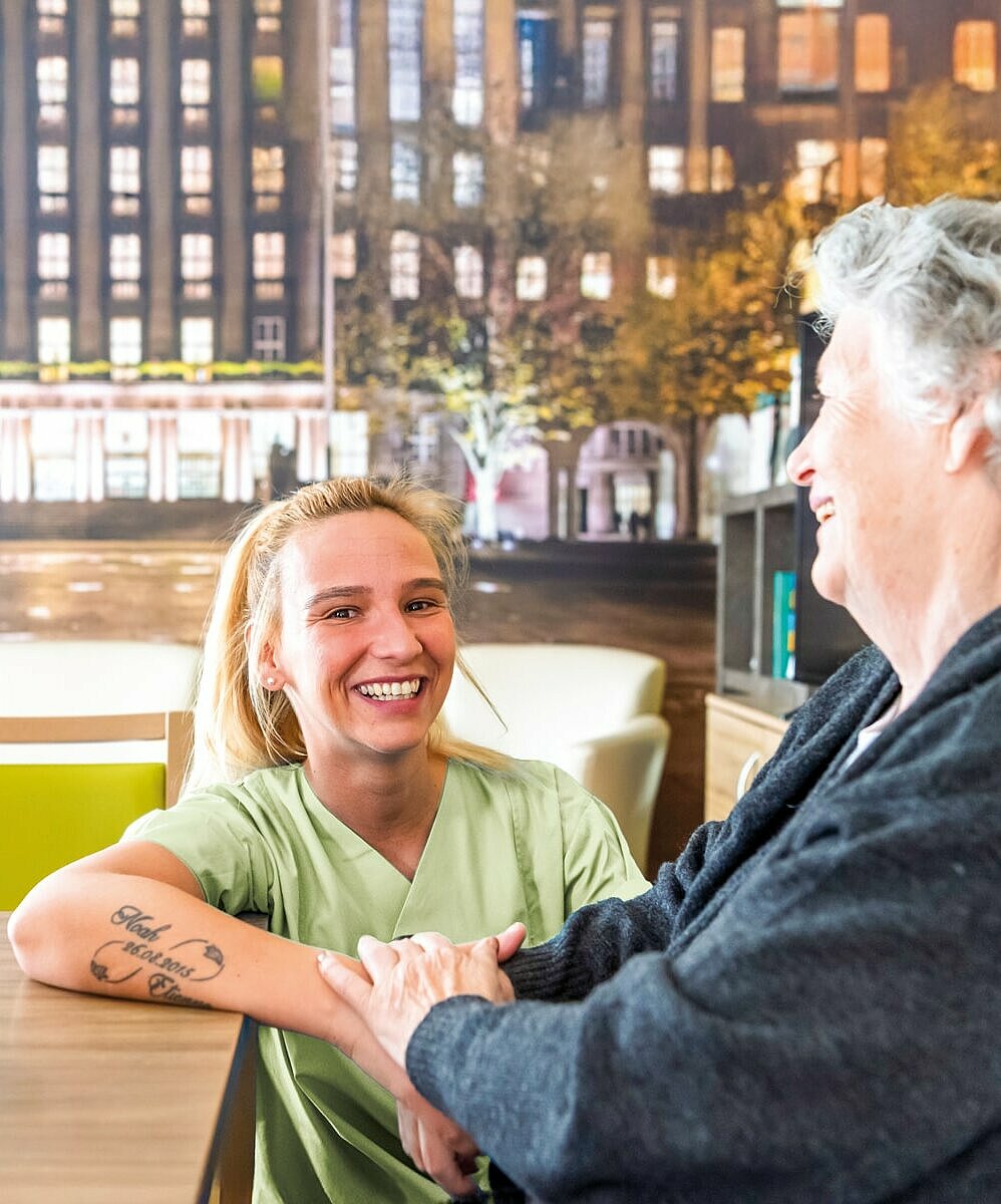 Eine lächelnde Pflegekraft hält die Hand einer fröhlichen älteren Frau, während sie an einem Tresen stehen.