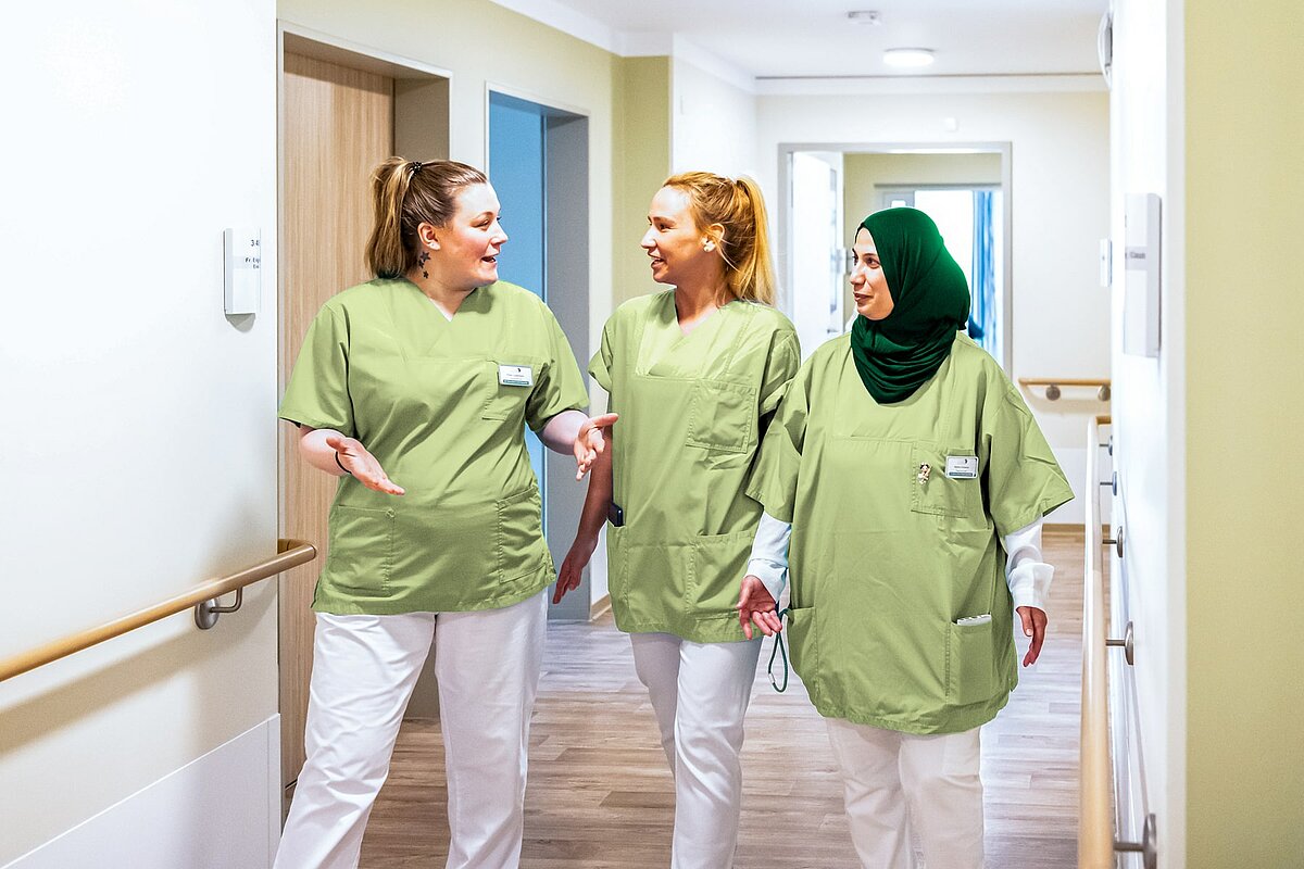 Drei lächelnde Pflegekräfte in grüner Arbeitskleidung und weißen Hosen unterhalten sich, während sie einen hellen Flur entlanggehen. Die Frau auf der rechten Seite trägt einen dunkelgrünen Hidschab.