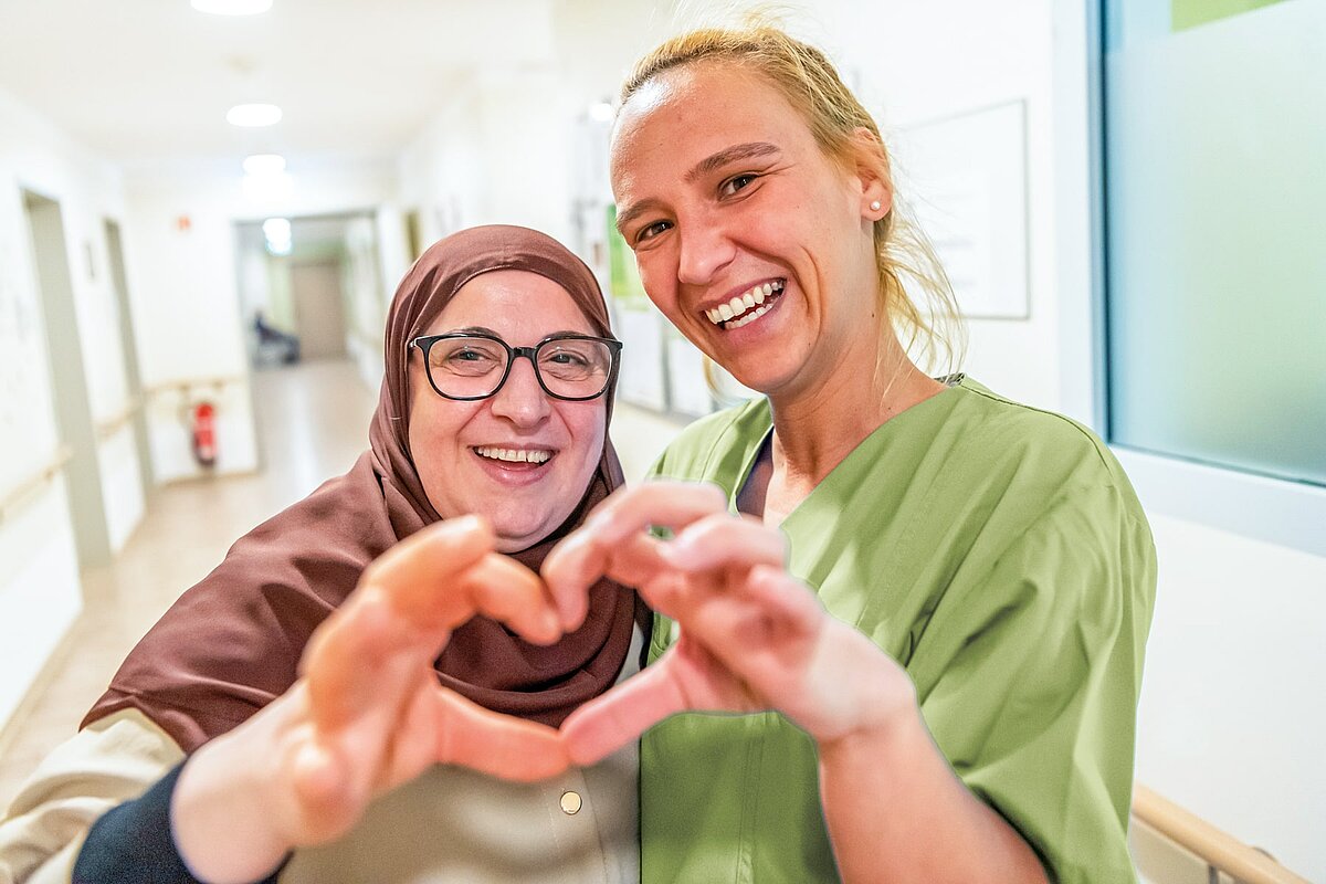 Eine lächelnde Frau mit Kopftuch und eine lächelnde junge Frau bilden mit ihren Händen ein Herz in einem Flur.