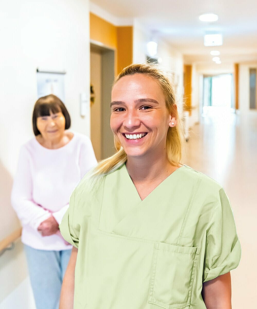 Eine lächelnde Pflegekraft steht im Vordergrund eines Pflegeheimflurs, während eine ältere Patientin im Hintergrund zu sehen ist.